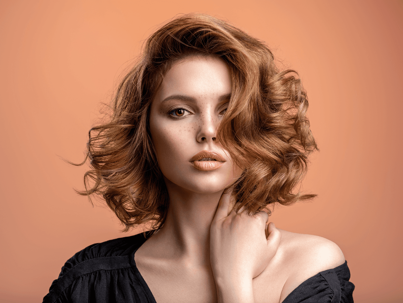 Woman with softly curled copper-toned hair, wearing a black top, against a warm peach background — representing seamless Aveda root regrowth colour at The Pamper Suite.