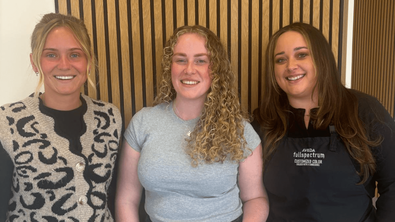 Three smiling women in salon, including a stylist in Aveda apron and a client with defined blonde curls. Represents curly hair maintenance at The Pamper Suite in Bexley.
