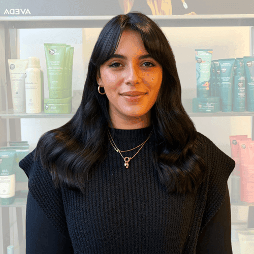 Jessica, professional hair stylist at The Pamper Suite in Bexley, smiling in front of Aveda product display.