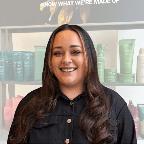Paige, expert colourist at The Pamper Suite in Bexley, smiling in front of Aveda haircare products.