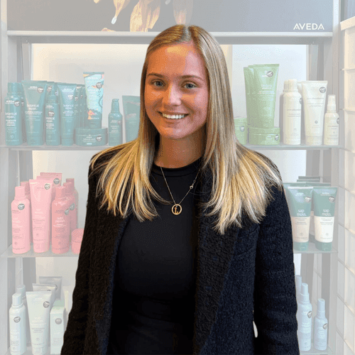 Sienna Sheppard, hair stylist at The Pamper Suite in Bexley, smiling in front of an Aveda product display.